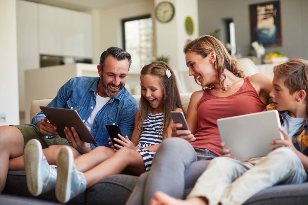 A happy family of four on the couch using tablets and phones together at home